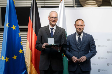 Zwei Männer im Anzug, einer erhält den Sächsischen Verdienstorden, vor EU-, Sachsen- und Deutschlandflagge.